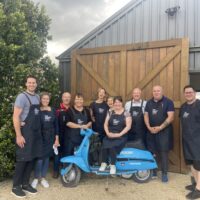 A cooking class group outside the barn with a blue Vespa